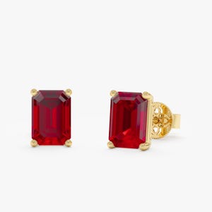 Op de afbeelding: Een paar oorstekers met rechthoekige, dieprode edelstenen gezet in goud. De gouden zetting heeft kleine, ronde accenten op elke hoek en een decoratief filigraanontwerp op de pin.