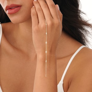 May include: A delicate gold hand chain featuring a series of small, oval-shaped gemstones. The chain extends from the wrist, across the back of the hand, and to the middle finger. The jewelry is worn against a light skin tone.