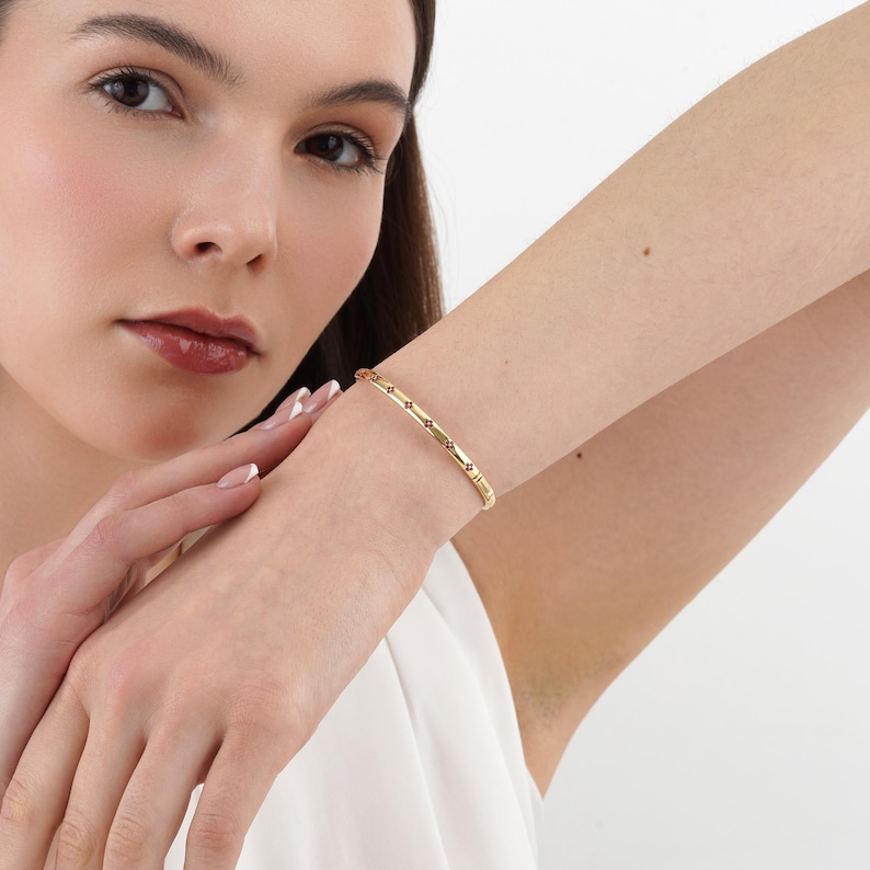 May include: A thin gold bangle bracelet featuring a repeating pattern of small, dark, four-pointed star or flower-shaped accents spaced evenly along the polished metal band, which rests securely around the wrist of the wearer.