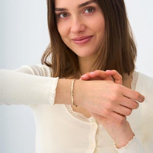 Puede incluir: Una pulsera dorada con un dise&ntilde;o delicado se lleva en la mu&ntilde;eca. La pulsera parece ser de metal y tiene un estilo sencillo y elegante. La persona lleva una blusa de color crema.