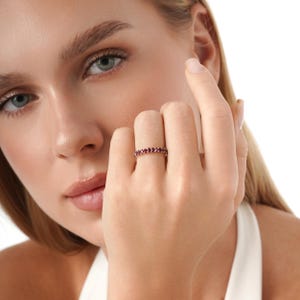 K&ouml;nnte beinhalten: Ein goldfarbener Ring mit einer durchgehenden Reihe kleiner, runder, roter Edelsteine. Der Ring wird an einem Finger getragen, wobei die Hand und ein Teil des Gesichts einer Person sichtbar sind. Der Hintergrund ist wei&szlig;.
