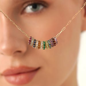 Könnte beinhalten: Eine goldfarbene Halskette mit einer Reihe von farbigen Edelsteinringen. Die Edelsteine sind in einem Regenbogenspektrum angeordnet, darunter Rot, Blau, Grün und Lila. Die Halskette wird um den Hals getragen.