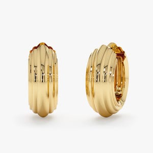 May include: Two gold-colored hoop earrings. Each earring has a ribbed, vertical design. The earrings are made of a shiny, reflective material. The earrings are displayed against a white background.