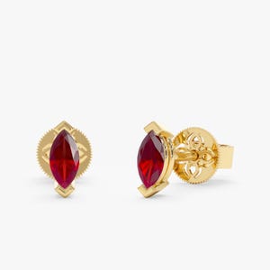May include: A pair of gold stud earrings with marquise-cut, deep red gemstones. The earrings have a geometric design around the gemstone and a decorative backing. The earrings are set against a white background.