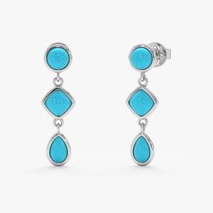 May include: A pair of silver drop earrings with turquoise-colored stones. Each earring features a round stone at the top, a square stone in the middle, and a teardrop-shaped stone at the bottom. The stones are set in a silver bezel.