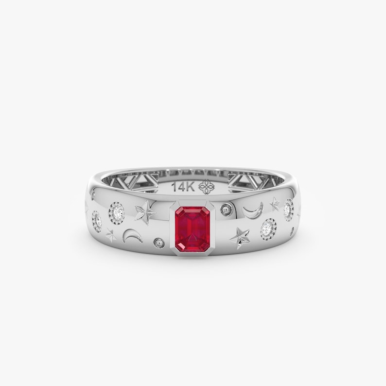 Op de afbeelding: Een zilverkleurige bandring met een centrale, smaragdgeslepen rode edelsteen, versierd met gegraveerde ster- en maansikkelmotieven en kleine ingelegde ronde steentjes. De binnenkant van de ring is duidelijk gestempeld met de tekst 14K.