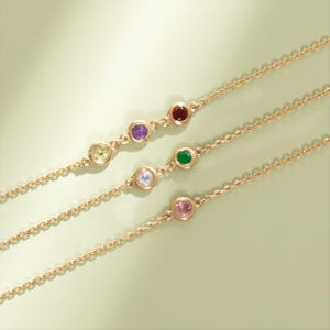 May include: Gold-coloured necklaces with colourful gemstones. Each necklace features a delicate chain and a series of round, bezel-set stones in various colours, including green, purple, and pink. The gemstones are evenly spaced along the chain.