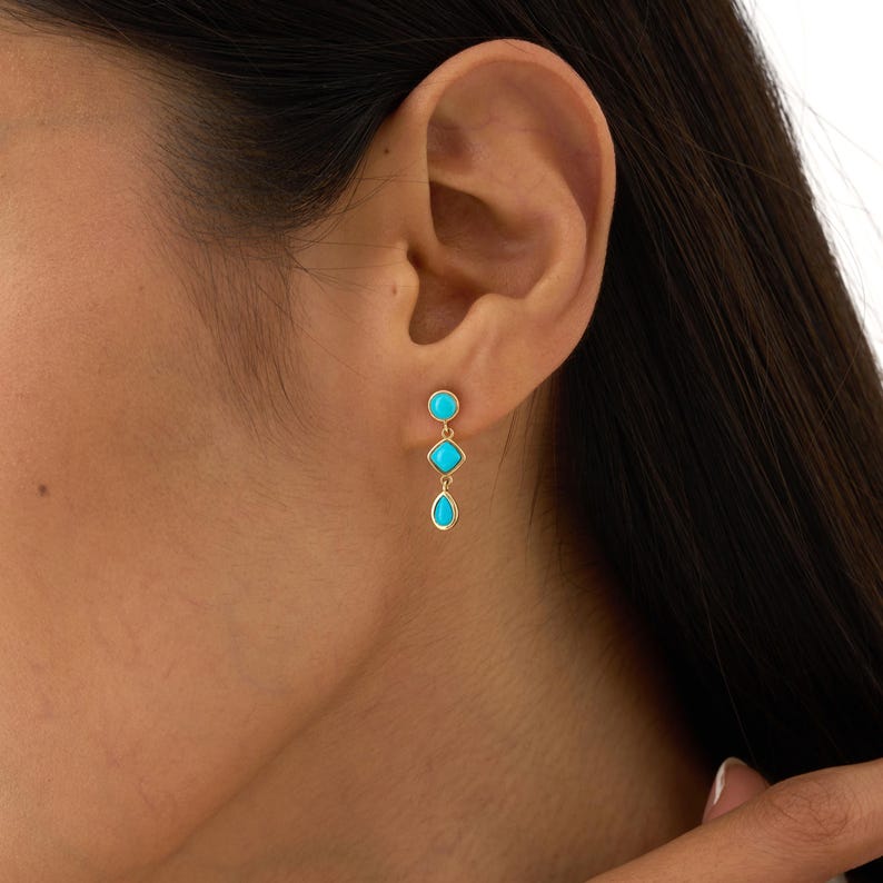 May include: Gold-toned drop earrings featuring three turquoise-colored stones. The top stone is round, the middle is square, and the bottom is teardrop-shaped. The earrings are dangling and are worn on the ear.