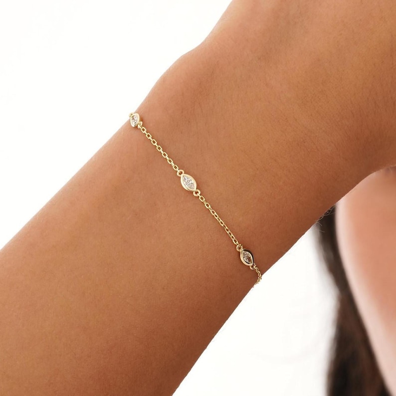 May include: A delicate gold bracelet featuring a chain with four bezel-set, marquise-shaped clear gemstones. The bracelet is shown on a person's arm, with the gemstones evenly spaced along the chain.