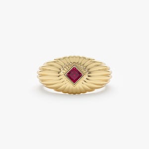 May include: A gold-colored ring with a central, square-cut, red gemstone. The ring has an oval shape with a fluted, fan-like design around the gemstone. The ring is set against a white background.
