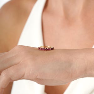 K&ouml;nnte beinhalten: Ein goldener Bandring mit oval geschliffenen, leuchtend roten Edelsteinen. Der Ring wird auf der Hand einer Person gezeigt, um das Design und die Farbe des Rings hervorzuheben. Der Ring ist ein Schmuckst&uuml;ck.