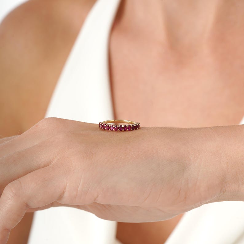 K&ouml;nnte beinhalten: Ein goldener Bandring mit oval geschliffenen, leuchtend roten Edelsteinen. Der Ring wird auf der Hand einer Person gezeigt, um das Design und die Farbe des Rings hervorzuheben. Der Ring ist ein Schmuckst&uuml;ck.