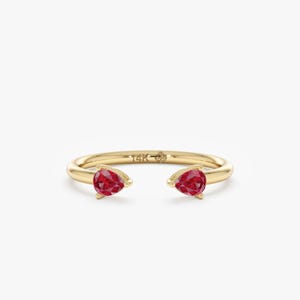 May include: A gold ring with two pear-shaped, red gemstones. The ring has an open design, with the gemstones facing opposite directions. The band is smooth and shiny, and the ring is set against a white background.