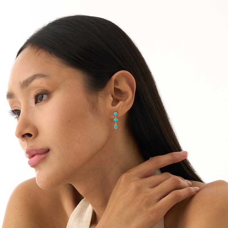 May include: A close-up of a woman wearing gold earrings with three turquoise-colored stones. The earrings are dangling and have a simple, elegant design. The woman has long, dark hair and is wearing a light-colored top.
