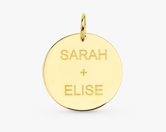 Puede incluir: Un colgante circular dorado con un lazo en la parte superior. El colgante presenta los nombres grabados "SARAH" y "ELISE" separados por un signo más. El fondo es de color claro sólido.