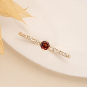 May include: A delicate gold ring featuring a single red gemstone surrounded by a row of smaller white gemstones.