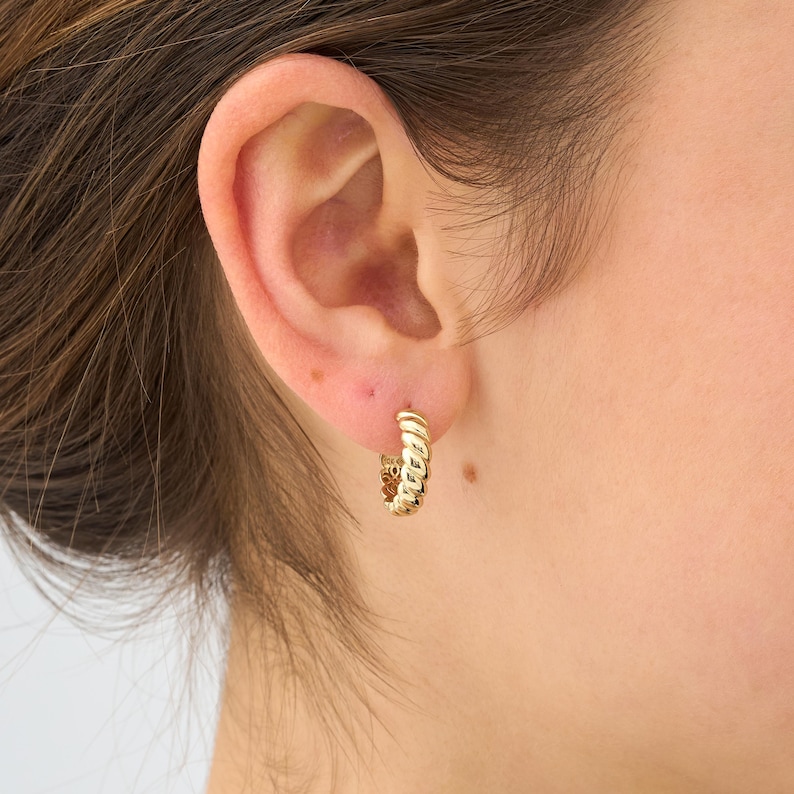 May include: A close-up of a gold-colored hoop earring. The earring has a textured, twisted design. The earring is worn on the ear of a person with light skin and brown hair.