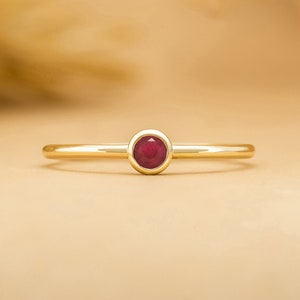 May include: A simple gold band ring with a small, round, bezel-set ruby gemstone.