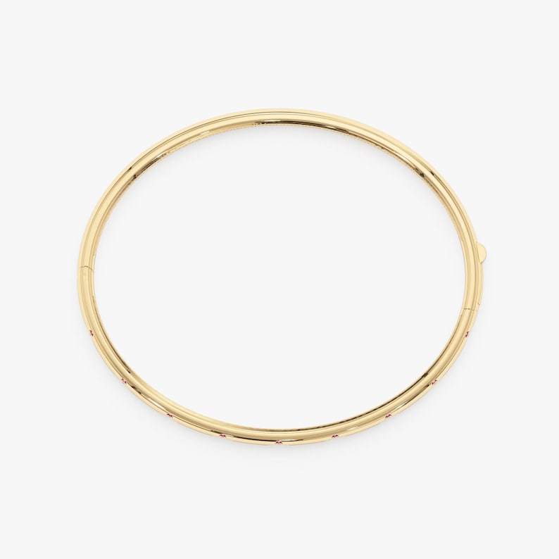 May include: A circular gold bangle bracelet featuring a polished metallic finish and a series of small, evenly spaced red gemstones embedded along the outer edge. The bracelet includes a visible hinged clasp mechanism on the side for opening.