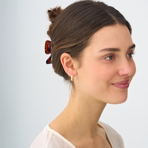 May include: A close-up shot of a person wearing gold hoop earrings. The earrings have a textured, twisted design. The person is wearing a light-colored top and has their hair pulled back with a brown hair clip.