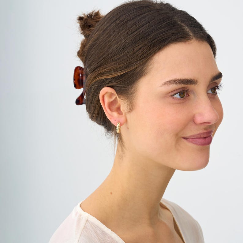 May include: A close-up shot of a person wearing gold hoop earrings. The earrings have a textured, twisted design. The person is wearing a light-colored top and has their hair pulled back with a brown hair clip.