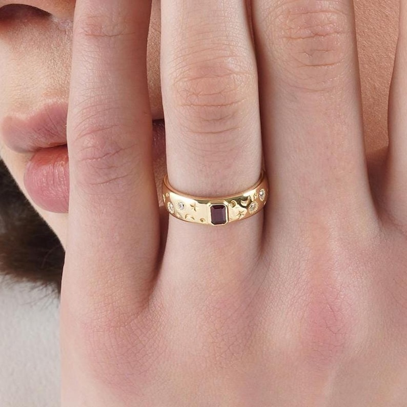 Op de afbeelding: Een gouden bandring met een centrale rechthoekige donkerrode edelsteen. Het oppervlak van de ring is gegraveerd met kleine ster- en maansikkelmotieven, afgewisseld met verschillende kleine, heldere, ronde, verzonken accentstenen.