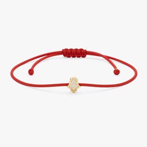 Könnte beinhalten: Rotes Armband mit einem goldfarbenen Hamsa-Hand-Anhänger, der mit kleinen, klaren Edelsteinen besetzt ist. Das Armband hat einen verstellbaren Schiebeknotenverschluss. Die Hamsa-Hand ist ein Symbol für Schutz und Glück.