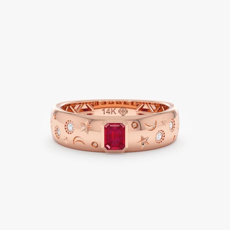 Op de afbeelding: Een 14K ros&eacute;gouden ring met een centrale rode edelsteen in emerald-slijpvorm. Het oppervlak is gegraveerd met hemelse motieven zoals sterren, maansikkels en kleine ronde accenten, ingezet met verschillende kleine ronde diamanten.