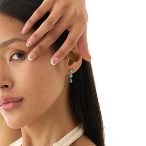May include: Gold-tone earrings with three turquoise-colored stones. The earrings are dangling and feature a round stone at the top, followed by a smaller round stone, and a teardrop-shaped stone at the bottom. The earrings are worn on a person's ear.