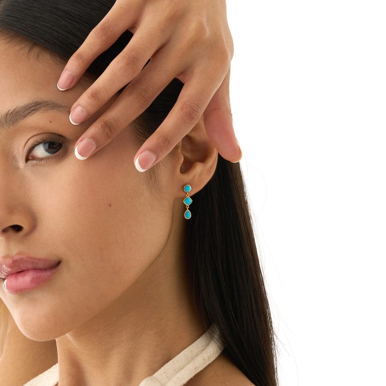 May include: Gold-tone earrings with three turquoise-colored stones. The earrings are dangling and feature a round stone at the top, followed by a smaller round stone, and a teardrop-shaped stone at the bottom. The earrings are worn on a person's ear.
