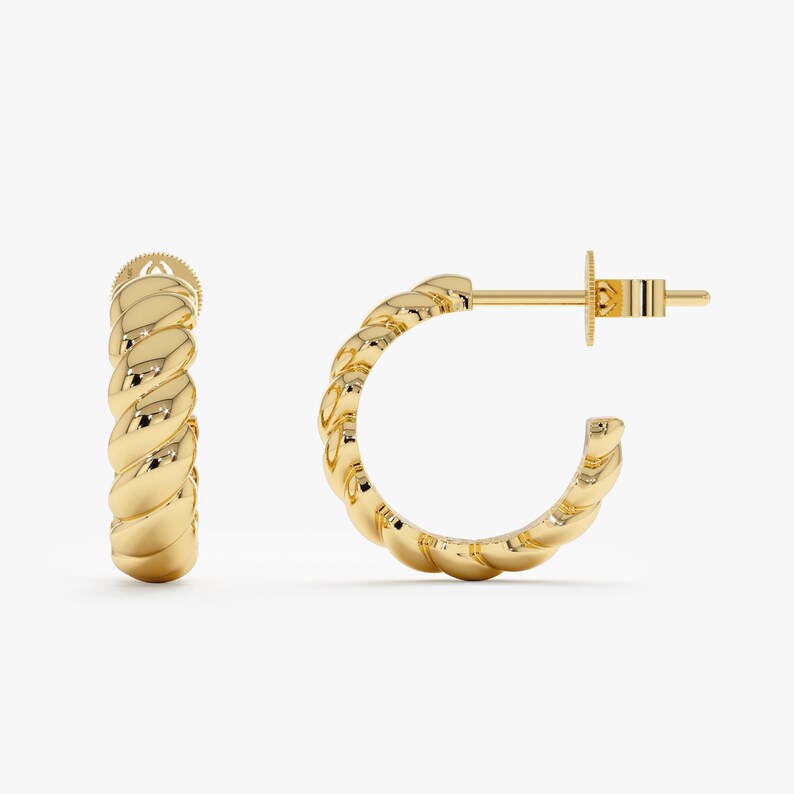 May include: A pair of gold-colored hoop earrings. Each earring has a twisted rope design. The earrings are displayed on a white background. The earrings have a post and butterfly clasp.