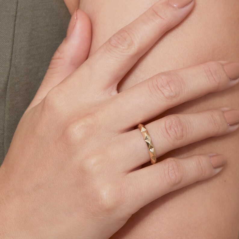 May include: A gold ring with a textured, triangular design. The ring is worn on a person's finger.