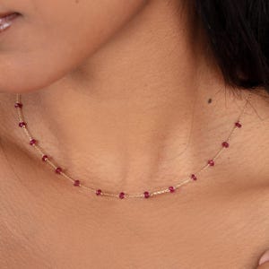 May include: A delicate gold necklace featuring small, round, ruby-colored gemstones. The gemstones are evenly spaced along the fine gold chain, creating a minimalist and elegant design. The necklace is shown on a person's neck.