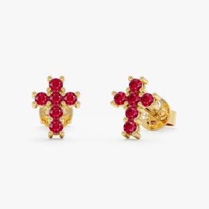 May include: A pair of stud earrings crafted from yellow gold, each featuring a cross-shaped design set with five round-cut vibrant red rubies. The earrings include secure post backings, showcasing a polished metal finish and faceted gemstones.