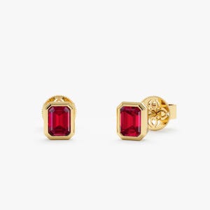 May include: A pair of stud earrings featuring emerald-cut red gemstones set in yellow gold bezels. The earrings have a polished gold finish and include detailed, intricate screw-back mechanisms visible on the side of one earring.