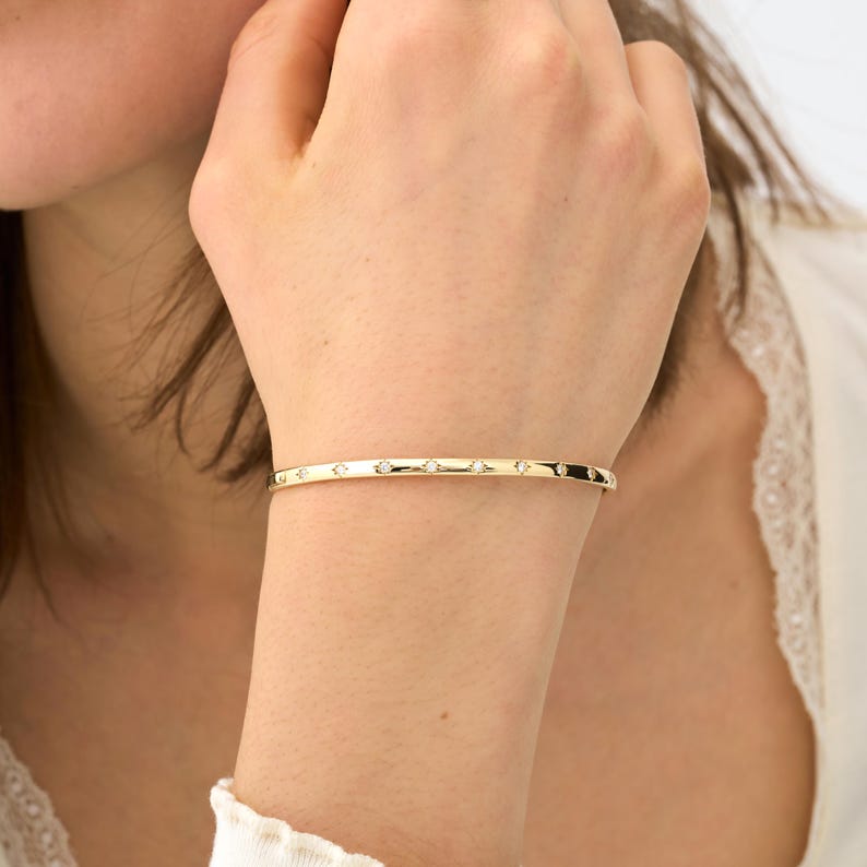 Puede incluir: Una pulsera de brazalete dorada con peque&ntilde;as gemas transparentes espaciadas uniformemente. La pulsera tiene un dise&ntilde;o simple y elegante. La pulsera se muestra en la mu&ntilde;eca de una persona.