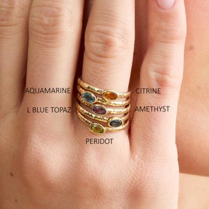 May include: A hand displaying several gold rings, each set with a different gemstone: aquamarine, citrine, amethyst, L blue topaz, and peridot. The rings have a textured band. Text on the image identifies each gemstone.