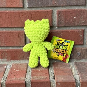 Peut inclure: Un jouet en peluche crocheté vert lime en forme de personne debout sur une surface en brique. Un paquet de bonbons Sour Patch Kids se trouve à côté du jouet.