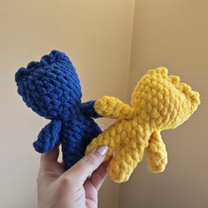 May include: Two crocheted stuffed toys, one blue and one yellow. The toys are holding hands and are made with a fluffy yarn.