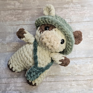 May include: A plush crochet stuffed animal featuring a cream-colored body with brown accents on the paws and face. The creature wears a sage green wide-brimmed hat and carries a matching sage green crossbody bag, with black plastic safety eyes.