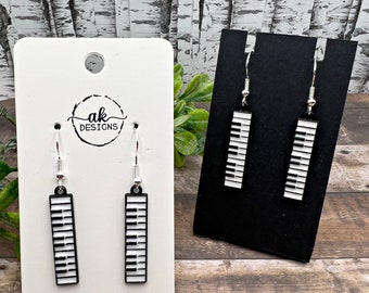 May include: A pair of silver dangle earrings with black and white piano key designs. The earrings are displayed on a white card with the text "ak DESIGNS".