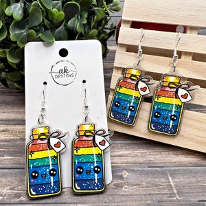 May include: Rainbow layered bottle charm earrings with a cute face and a small heart tag. The earrings are on a white card with the text "ak DESIGNS".
