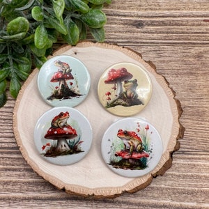 May include: Four round button pins with a white background and a colorful illustration of a frog sitting on a red and white mushroom. The mushrooms have white spots and the frogs are green with brown spots.