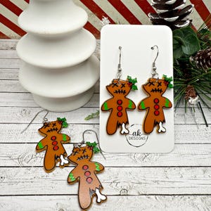 May include: A pair of dangle earrings featuring gingerbread people with stitched-up mouths, red buttons, and bone-like legs. The gingerbread people are wearing green holly leaves. The earrings are on a white card with the text "ak Designs".