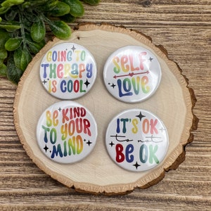 May include: Four round buttons with rainbow coloured text and designs. The buttons say "Going to Therapy is Cool", "Self Love", "Be Kind to Your Mind", and "It's Ok to Not Be Ok".