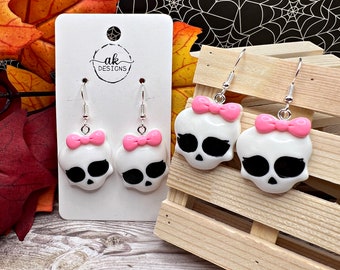 May include: White skull earrings with black eye sockets and a pink bow. The earrings are on a white card with the text "ak designs". The background includes fall leaves and a spiderweb.