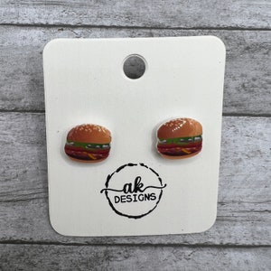 May include: A pair of stud earrings featuring a miniature hamburger design. The earrings are white with a brown bun, green lettuce, red tomato, and yellow cheese.