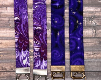 May include: Four fabric wrist keychains in shades of purple. Two feature a marbled design, and two have a starry night pattern with gold stars. Each keychain has a metal ring and a matching metal clasp.