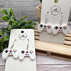 May include: White game controller earrings with red hearts and colorful buttons. The earrings are on a white card with the text "ak designs".