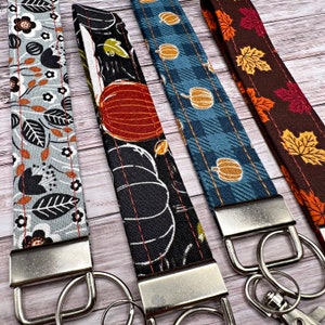 May include: Four fabric wrist keychains with fall-themed patterns. The patterns include floral, pumpkins, and fall leaves in shades of gray, black, blue, orange, and brown. Each keychain has a silver-colored metal clasp and key ring.